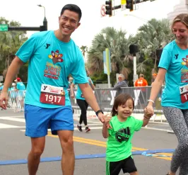 Outdoor running race with a family of three smiling at the finish line. The tall dad wears all blue, holding hands with his toddle and mom. Mom is wearing a matching Turkey Gobble and pushing the empty stroller.