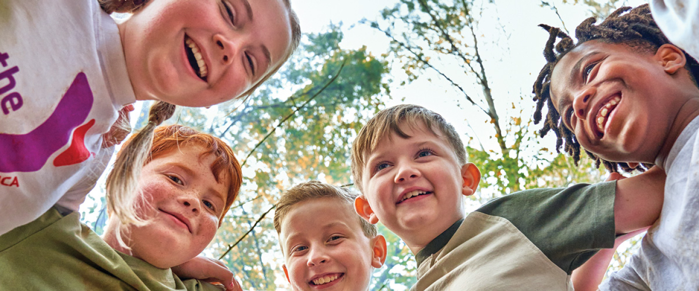 Five kids huddling, smiling, looking down at the camera, lens is pointing up at their faces. Trees background of the photo.