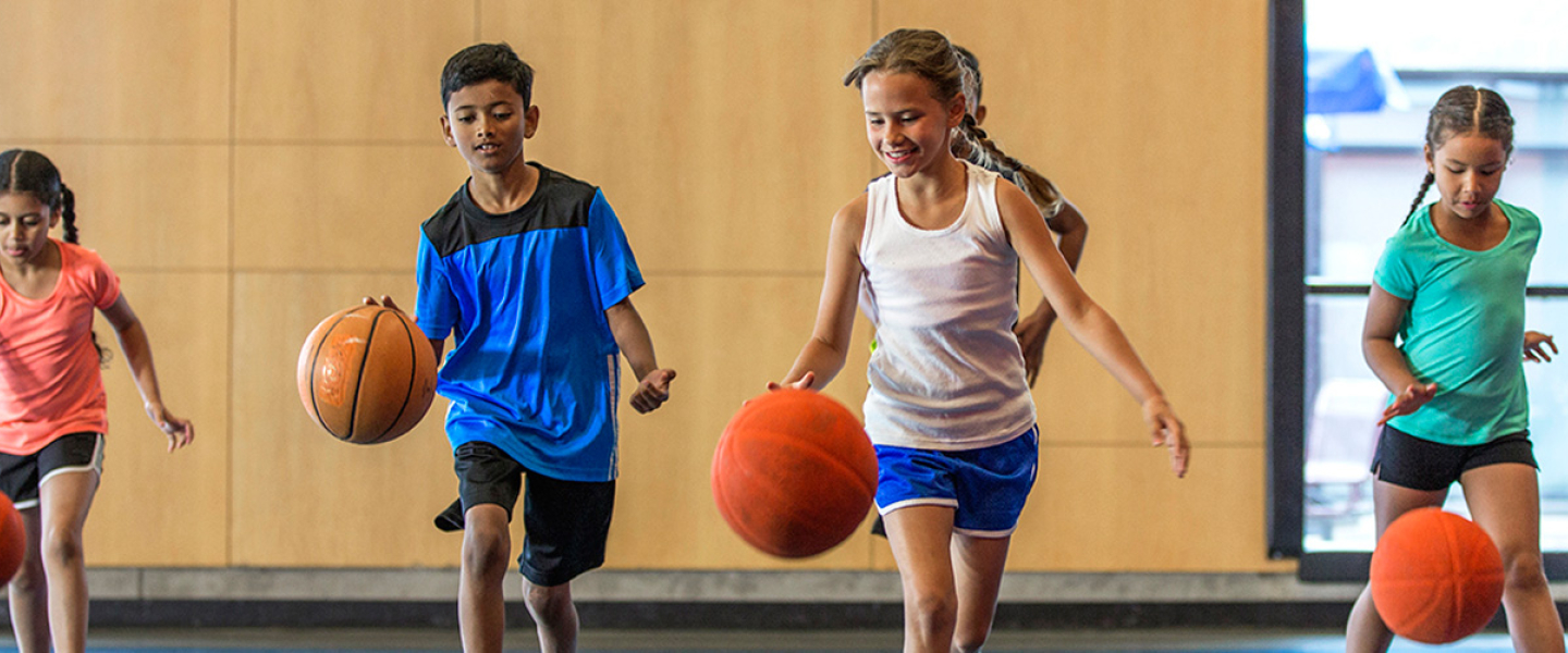 Four school age kids bouncing basketballs in indoor basketball gym.