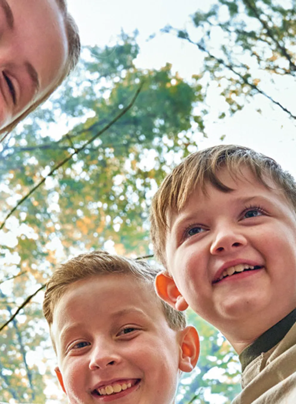 Five kids huddling, smiling, looking down at the camera, lens is pointing up at their faces. Trees background of the photo.