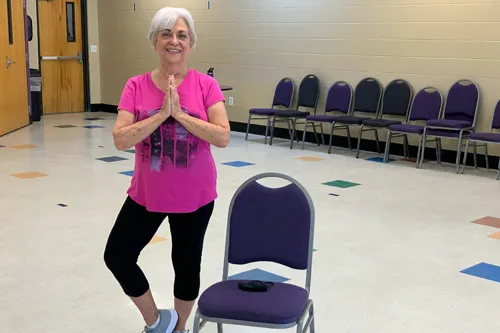 active older adult stands in tree pose by a chair at the ymca