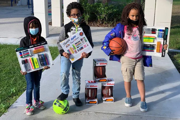 three kids wearing face coverings outdoors showing their new writing utensil kits and sports gear