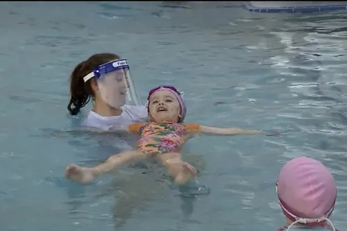 Swim instructor wearing face shield assisting child float on back in pool