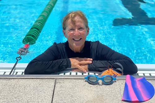 woman wearing rash guard resting her arms on ledge of pool. Visor and goggles on pool deck.