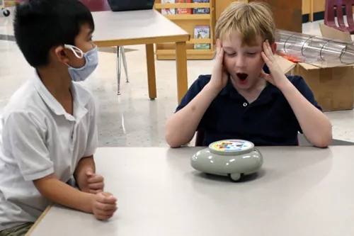 two kids at a table with a circular robot. one child showing a shocked emotion with hands on face and other child wears a face covering looking at other kid.