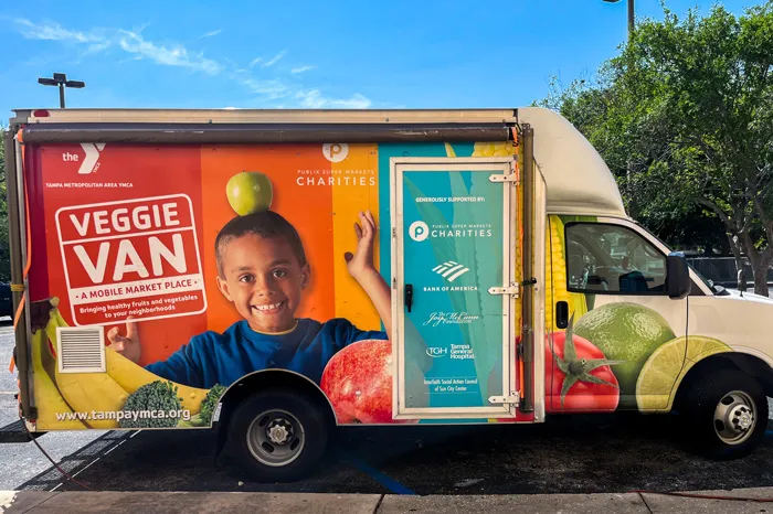 The Tampa YMCA Veggie Van wrapped with graphic logo, a child smiling balancing and apple on head, a colorful background and images of fruits and vegetables. 