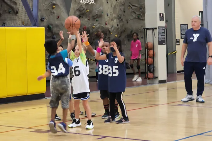 Youth co-ed YMCA basketball teams playing in an indoor court, under the basket. A coach wearing a navy blue shirt with a white Y logo watches the team play. Yellow wall padding and climbing rock wall are in the background.