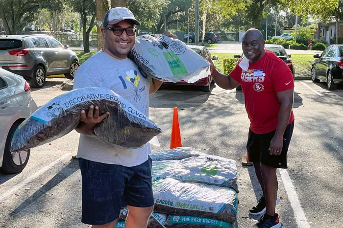 Two men in parking lot with bags of mulch smile at the camera during a volunteer day at the YMCA.
