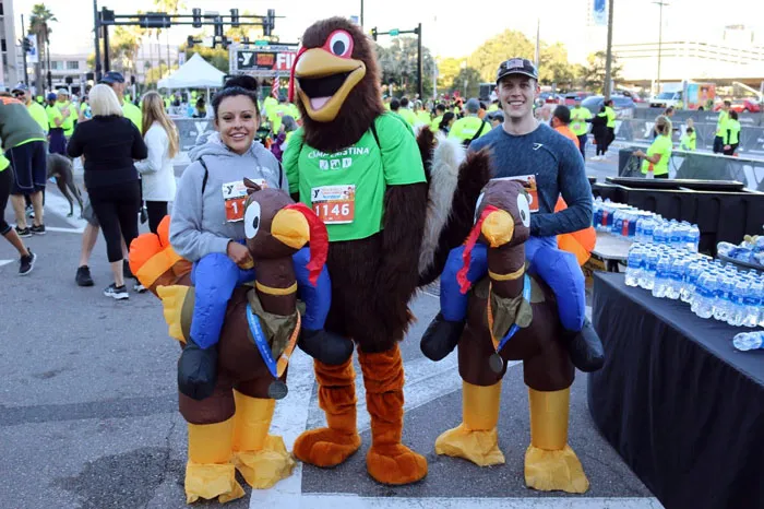 The Tampa YMCA Turkey Gobble mascot poses with two race finishers, dressed in turkey costumes. The image is outdoors, and the background is filled with people celebrating the end of the race, and black table cloth with water bottles.