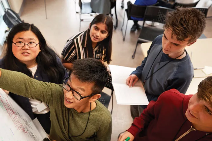 Five teens working together on a group project at a whiteboard on the wall.