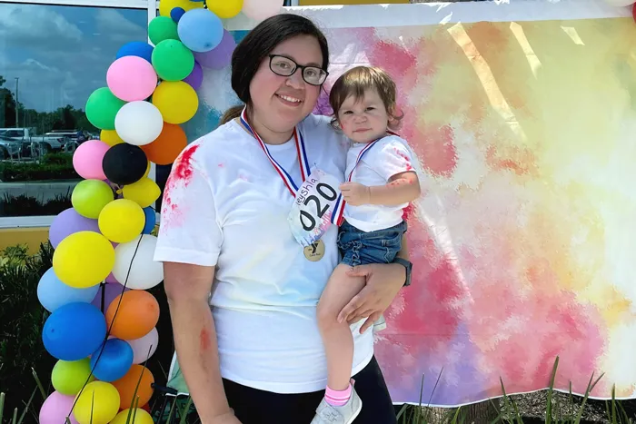 A mother holds toddler in front of colorful balloon arch and step and repeat backdrop outdoors at Spurlino Family YMCA color run fundraiser.