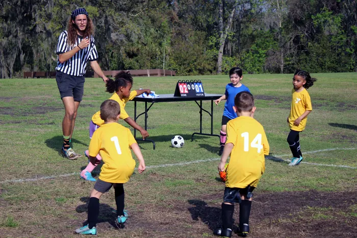 A volunteer referee calls a youth soccer game. Outdoors on a sunny day, four young soccer players wear yellow t-shirts and one wears blue.