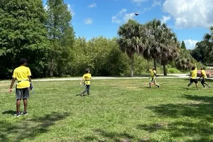 Five teens wearing yellow shirts play football outdoors. Scenery includes green grass, large trees and palm trees, a blue sky with white clouds.
