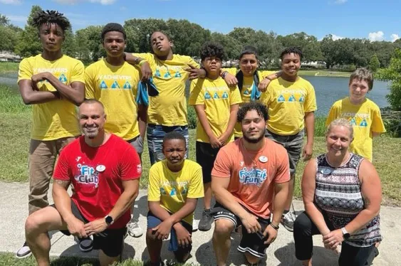 Eight Tampa Y Teen Achievers summer 2023 campers wearing matching yellow shirts pose with three staff members in an outdoor group photo. Background includes green grass, a large pond, green trees and blue skies.