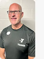 Male personal trainer wearing a black YMCA trainer t-shirt in front of a gray wall.