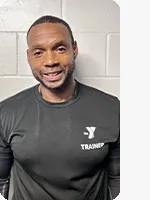 Male personal trainer wearing a black YMCA trainer t-shirt in front of a gray wall.