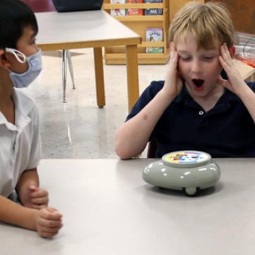 two kids at a table with a circular robot. one child showing a shocked emotion with hands on face and other child wears a face covering looking at other kid.