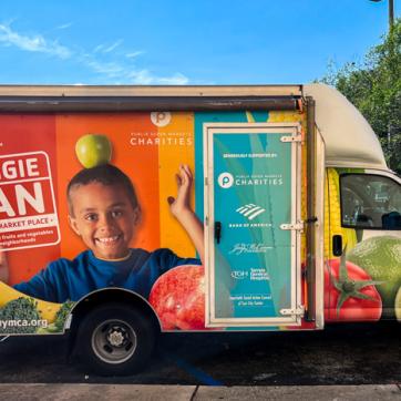 The Tampa YMCA Veggie Van wrapped with graphic logo, a child smiling balancing and apple on head, a colorful background and images of fruits and vegetables. 