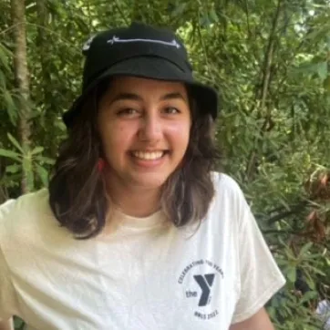 Teen wearing a white YMCA t-shirt and black bucket hat. Green shrubs and trees in the background.