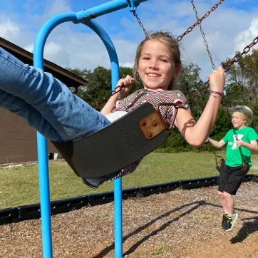 two kids on outdoor swings. girl wearing jeans in the center image, boy wearing green shirt swings in background.