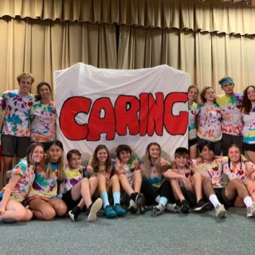 Group of teens wearing tie dye shirts gathered with a large DIY Caring sign. Sign has a white background and large, filled in red letters for "Caring."