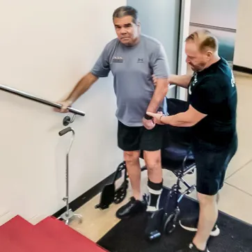 YMCA personal trainer and Y member regaining strength after a stroke, stand at the bottom of a staircase. The member stands in front of his wheelchair and holds onto the railing, while the trainer supports him.
