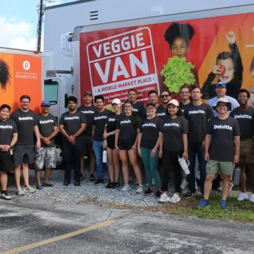 A group of 20 volunteers from Deloitte, wearing black t-shirts with their white logo, stand for a photo in front of Tampa YMCA's Veggie Van. The scene is outdoors on a sunny day. The group stands in a parking lot and patches of rocks and grass. 