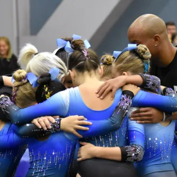 North Brandon Top Flight gymnastics team huddles with their coach at the 2023 YMCA National Gymnastics Championship. The coach wears a black short sleeve shirt and youth gymnasts wear blue and black gradient performance leotards with vertical line sparkles.