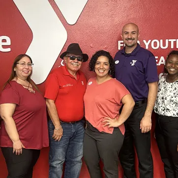 7 YMCA employees stand together in front of Y logo on the wall behind them