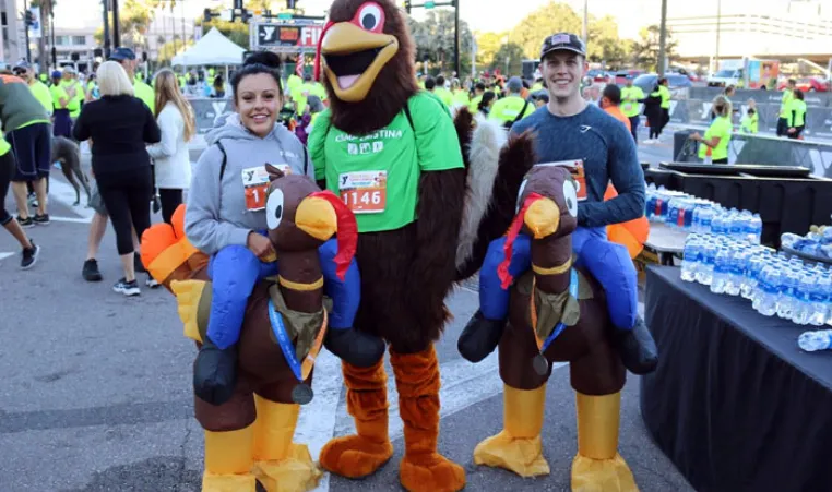 The Tampa YMCA Turkey Gobble mascot poses with two race finishers, dressed in turkey costumes. The image is outdoors, and the background is filled with people celebrating the end of the race, and black table cloth with water bottles.