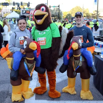 The Tampa YMCA Turkey Gobble mascot poses with two race finishers, dressed in turkey costumes. The image is outdoors, and the background is filled with people celebrating the end of the race, and black table cloth with water bottles.