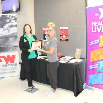 photo taken with two women - one receiving a Fire Watch certificate from the other in between two banners - one for Fire Watch and one for YMCA Healthy Living initiatives.