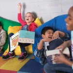 Four kids sitting on colorful bean bags, laughing and smiling with books in their hands.