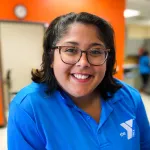 Headshot of female staff smiling at the camera in the YMCA Welcome Center. Staff is wearing a blue polo shirt with a Tampa Y logo, orange wall in background.