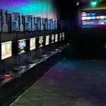 Dark computer room with colorful lights. Computers are mounted on the wall, with monitors underneath. Person standing at computer on the right.