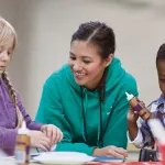 Two school age children making crafts with smiling counselor.